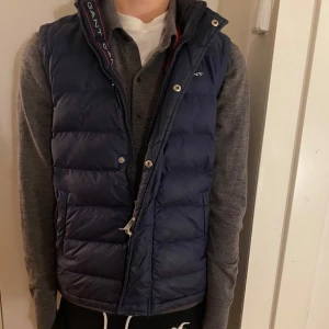 Mörkblå dunväst från GANT - Säljer en snygg mörkblå dunväst från GANT i mycket bra skick. Västen har en dragkedja och tryckknappar framtill, samt en röd insida som ger en cool kontrast. Modellen är 170cm lång. Hör av er vid frågor och funderingar!😁