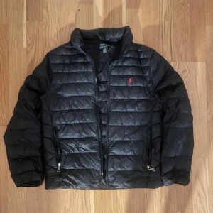 Svart pufferjacka från Polo Ralph Lauren - Säljer en snygg svart pufferjacka från Polo Ralph Lauren i mycket bra skick. Jackan har en quiltad design med dragkedja och det klassiska röda logotypbroderiet på bröstet. Perfekt för höst och vinter, håller dig varm och stilren. Passar både till vardags och lite finare tillfällen.