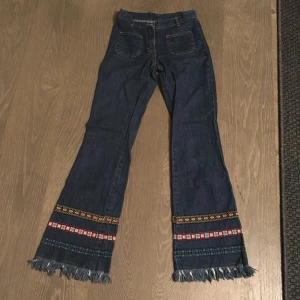 Unika jeans med broderade detaljer - Säljer ett par coola jeans med broderade mönster i rött, orange och blått längst ner på benen. De har en bootcut-stil och fransar vid bensluten. Perfekta för en bohemisk look! Jeansen är i bra skick och har praktiska fickor både fram. Midjemått ca 34cm innebenslängd ca 78cm