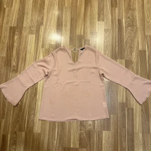 Rosa blus med ringdetalj - En jättefin rosa topp från ALLY BELL i storlek M med en unik ringdetalj vid nacken. Blusen har långa ärmar med en lätt utsvängd design. Perfekt för både vardag och festliga tillfällen. Oanvänd!!