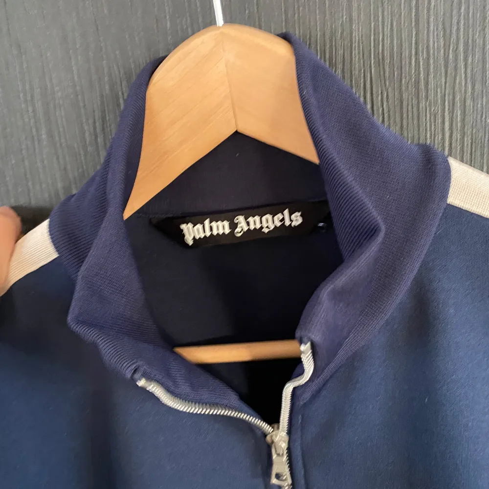 Säljer en snygg mörkblå track jacket från Palm Angels. Den har en dragkedja framtill och vita ränder längs ärmarna. Perfekt för en casual look eller träning. Jackan är i mycket bra skick och har en broderad logga på bröstet.. Jackor.