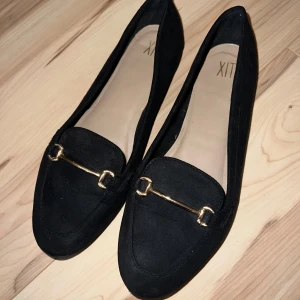 Svarta loafers med gulddetalj - Snygga svarta loafers från XIT i storlek 38. De har en elegant gulddetalj framtill som ger en lyxig touch. Perfekta för både vardag och fest. Skorna är i bra skick och redo att användas direkt. Passar 39. Inte fri frakt.