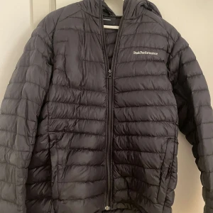 Svart dunjacka från Peak Performance - Säljer en svart dunjacka från Peak Performance i mycket bra skick. Jackan har dragkedja framtill. Perfekt för kalla höst- och vinterdagar. Den har en normal passform och långa ärmar. Huvan ger extra skydd mot väder och vind. Passar både till vardags och utomhusaktiviteter. Den är i storlek L. 