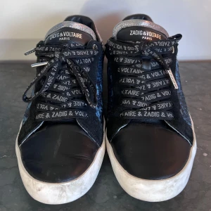 Svarta sneakers från Zadig & Voltaire - Snygga svarta sneakers från Zadig & Voltaire i skinn. De har coola snören med märkeslogga och en vit sula som ger en skön kontrast. Perfekta för en casual look med en touch av stil. Använda men i bra skick.                                                      Nypris: 2000kr 