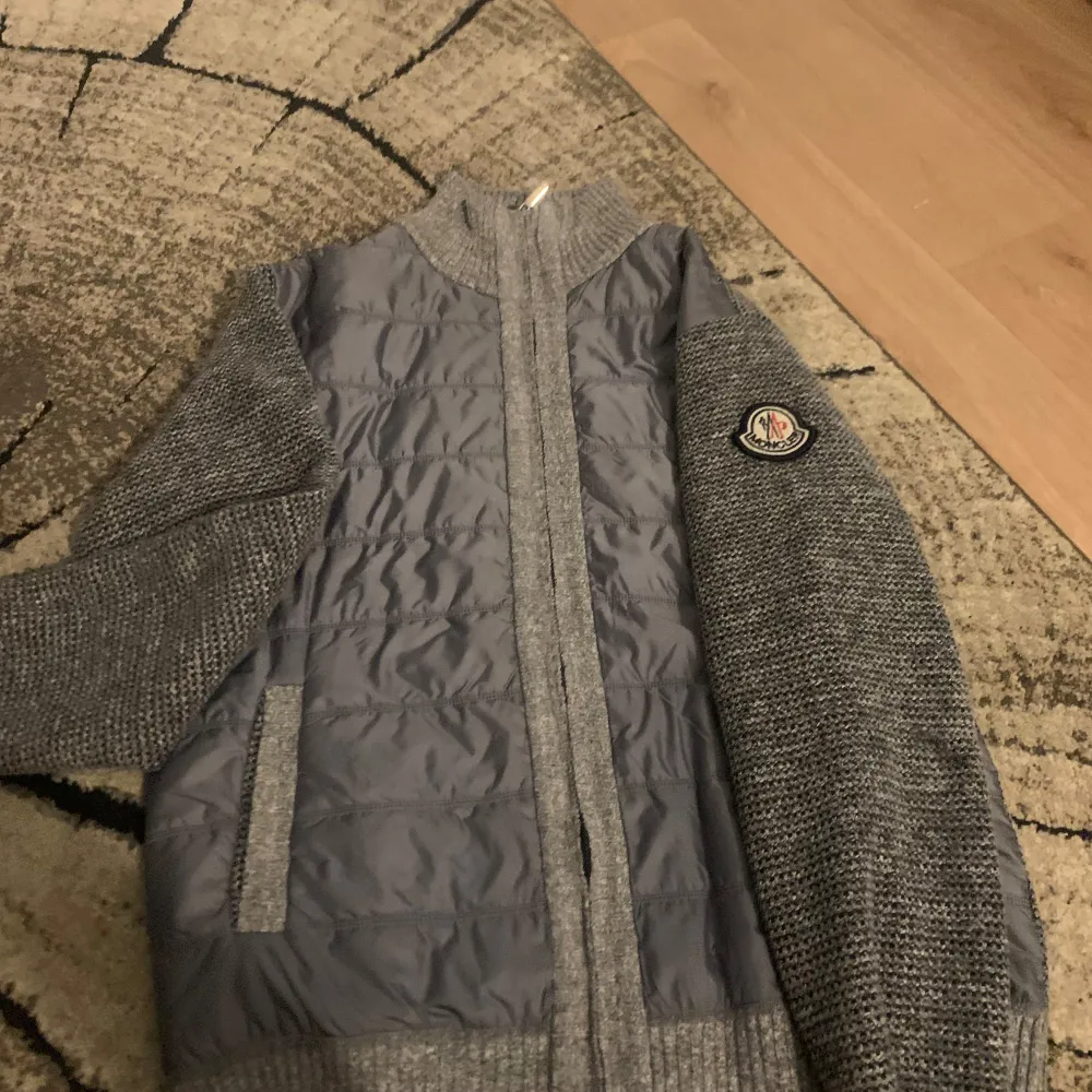 Säljer en snygg grå stickad jacka från Moncler. Den har quiltade detaljer på framsidan och ärmarna är långärmade och stickade. Perfekt för höst och vår! Jackan har en dragkedja framtill och Moncler-loggan på ärmen. Superbekväm och stilren!. Jackor.