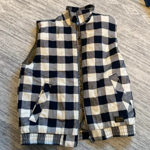 Rutig väst från Vailent - Säljer en snygg rutig väst från Vailent i svart och vit. Den har en dragkedja framtill och är quiltad på insidan för extra värme. Perfekt för höst och vår när du vill ha en stilren look. Västen är i mycket bra skick.