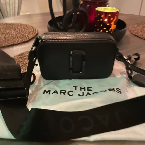 Svart axelväska från Marc Jacobs - Snygg svart axelväska från Marc Jacobs i skinn. Väskan har en stilren design med en stor metallspänne på framsidan och en justerbar axelrem. Perfekt för både vardag och fest!