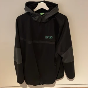 Svart fleece från Hugo Boss - Säljer en snygg svart fleece från Hugo Boss med grå detaljer och dragkedja. Perfekt för höst och vår, med en sportig look och huva. Fleecen har långa ärmar och är i mycket bra skick. Passar perfekt för både vardag och träning.
