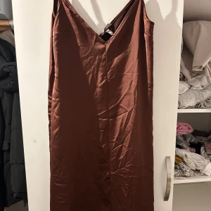 Brun axelbandslös klänning - Säljer en elegant brun axelbandslös klänning i silke material. Perfekt för festliga tillfällen med sin stilrena design och v-ringade framdel. Klänningen är i mycket bra skick och passar perfekt för höst och vinter.