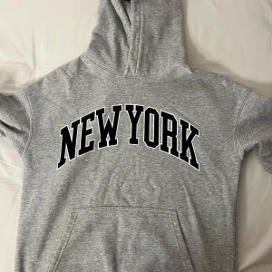 Grå hoodie med New York-tryck - Säljer en grå hoodie med stort 'New York'-tryck på framsidan. Den är supermjuk och perfekt för kyliga dagar. Har en klassisk känguruficka och en bekväm huva. Perfekt för en avslappnad stil eller när du vill mysa hemma. Passar till allt!