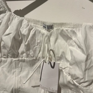 Vit topp från Bershka - Säljer en superfin vit blus/topp från Bershka i storlek M. Den har en smockad detalj på baksidan och knytning framtill. Perfekt för sommardagar eller en avslappnad vårlook. Toppen är i nytt skick, aldrig använt och har en luftig passform med långa ärmar. ORD PRIS 230kr