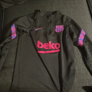 Svart Nike träningströja med Barcelona-logga - Säljer en svart långärmad träningströja från Nike med FC Barcelona-logga och Beko-tryck i rosa. Tröjan har en halv dragkedja framtill och coola mönstrade detaljer i rosa och blått på ärmarna. Perfekt för fotbollsträning eller som en snygg vardagströja!  Storlek 158-165