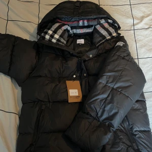 Svart dunjacka från Burberry - Säljer en snygg svart dunjacka från Burberry i nytt skick. Jackan har en quiltad design med dragkedja och knappar framtill. Den har en avtagbar huva med ett klassiskt rutigt mönster på insidan. Perfekt för kalla vinterdagar! 🖤