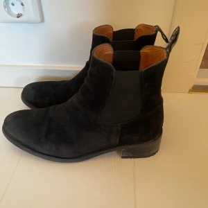 Svarta mocka Chelsea boots från Tiger of Sweden  - Säljer ett par stilrena svarta Chelsea boots i mocka från Tiger of Sweden. De har en klassisk design med en liten klack. Perfekta för både vardag. Använda men i mycket bra skick. Har adderat en gummisula på klacken hos skomakare. Storlek 40, passar 39-40.
