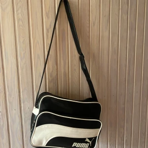 Träningsväska från Puma - Säljer en snygg svart och vit axelväska från Puma. Väskan har en justerbar axelrem och flera fack med dragkedja för enkel förvaring. Perfekt för vardagsbruk eller träning. Den är i bra skick och redo för nya äventyr!