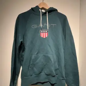 Säljer en snygg grön hoodie från GANT i storlek M. Den har en klassisk design med GANT-loggan och ett coolt emblem på bröstet. Perfekt för höst och vinter med sin mysiga känsla och långa ärmar. Huvan har justerbara snören och det finns en praktisk magficka framtill.