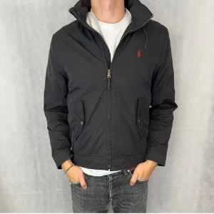 Svart vindjacka från Ralph Lauren - Snygg svart vindjacka från Ralph Lauren med dragkedja och huva. Jackan har en liten röd logga på bröstet och praktiska fickor med knappar. Perfekt för höst och vår, och passar till både vardag och fest. Den är i mycket bra skick och redo för nya äventyr! Det går att sänka priset vid snabb affär! Det går också att välja om du vill ha huva eller inte, samt så kan du göra så att armarna sitter tajtare. 