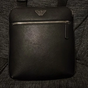 Svart axelväska från Armani - Snygg svart axelväska från Armani i skinn med en stilren design. Väskan har en praktisk dragkedja framtill och ett diskret märke i silver. Perfekt för både vardag och fest!