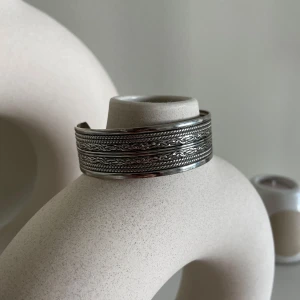 Silverfärgat armband med detaljerat mönster - Säljer ett snyggt silverfärgat armband med ett detaljerat och intrikat mönster. Armbandet är öppet och justerbart, vilket gör det lätt att ta på och av. Perfekt för att ge en stilren touch till vilken outfit som helst. Passar både till vardags och fest!
