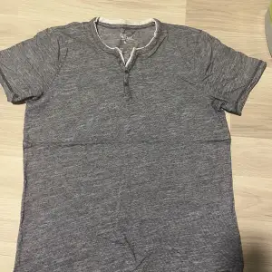 Säljer en snygg grå t-shirt från Jean Pascale i storlek M. Den har en knappdetalj vid halsen och är kortärmad. Perfekt för en avslappnad stil och passar bra till både jeans och shorts. T-shirten är i bra skick och redo för nya äventyr!