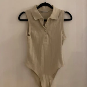 Beige ribbad ärmlös body från Zara - Säljer en stilren beige ribbad body från Zara. Den är ärmlös med en klassisk krage och knappar framtill. Perfekt för både vardag och fest! 🥰
