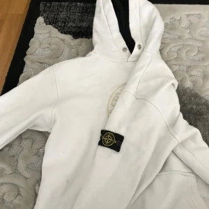 Vit hoodie från Stone Island - Säljer en snygg vit hoodie från Stone Island med deras ikoniska logga broderad på bröstet. Tröjan har en klassisk passform och är perfekt för både vardag och chill. Den har en justerbar huva och ribbade muddar för extra komfort. Perfekt för höst och vår!