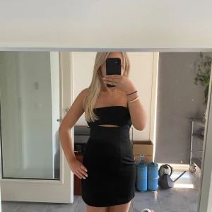 Svart axelbandslös klänning - Snygg svart axelbandslös klänning med en stilren design och en liten utskärning framtill. Perfekt för festliga tillfällen! Klänningen är tight och kort, vilket ger en elegant och modern look.