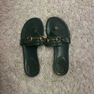 Gröna sandaler från Gucci - Snygga gröna sandaler från Gucci i bra skick. De har en elegant gulddetalj på ovansidan och är perfekta för sommaren. Sandalerna är tillverkade i skinn med en texturerad yta som ger en lyxig känsla.