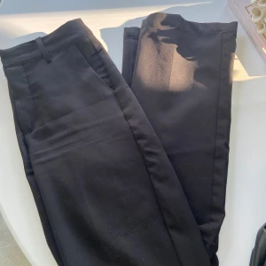 Svarta kostymbyxor - Säljer ett par svarta kostymbyxor i bra skick. De är högmidjade och har en klassisk straight passform. Perfekta för både kontoret och festligare tillfällen. Byxorna har en enkel design utan mönster och är gjorda i ett bekvämt material.