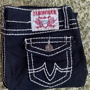 Svarta jeans med kontrastsömmar - Snygga svarta jeans från Fashion Denim med vita kontrastsömmar som verkligen poppar! De har en cool fickdesign med knappdetalj och ett märke på baksidan. Perfekta för en casual look eller en utekväll. Passar bra till både sneakers och boots. De likar True Religions och har mjuk tyg istället för jeans material.