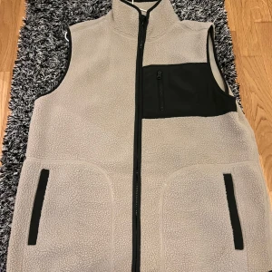 Beige fleeceväst med dragkedja - Säljer en mysig beige fleeceväst med grön dragkedja och praktiska fickor. Perfekt för kyliga dagar och passar bra till lager-på-lager. Västen har en hög krage och en bröstficka med dragkedja för extra förvaring. 
