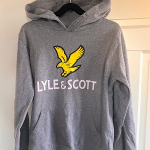 Grå hoodie från Lyle & Scott - Säljer en snygg grå hoodie från Lyle & Scott i storlek 15/16 år. Den har en stor gul logga med en fågel på framsidan och är supermjuk och bekväm. Perfekt för både skola och chill hemma. Huvan är skön och ger extra värme under kyliga dagar.