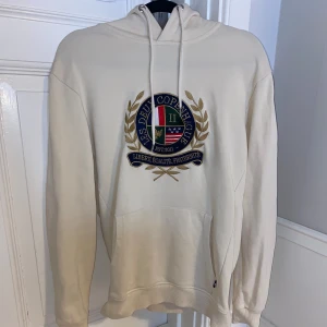 Beige hoodie från Les Deux - Säljer en snygg beige hoodie från Les Deux med ett coolt emblem på bröstet. Den har en klassisk känguruficka och justerbar huva. Perfekt för både höst och vår, och passar till nästan allt. Skön och stilren design!