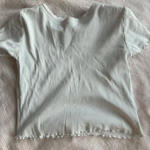 Vit kortärmad tröja från Zara - Säljer en enkel och stilren vit topp från Zara i storlek 134-140. Den är kortärmad och har en lätt ribbad struktur med vågig kant nedtill. Perfekt för vardagsbruk eller att matcha med en snygg jacka. Toppen är i bra skick och redo för en ny ägare!
