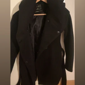 Svart kappa från New yorker - Säljer en elegant svart kappa från New yorker i mycket bra skick. Den har en stilren design med knappar framtill och en skön passform. Perfekt för höst och vinter, håller dig varm och snygg. Passar både till vardags och fest!