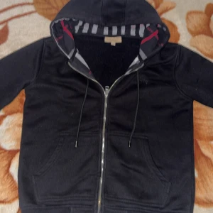 Svart hoodie från Burberry - Säljer en snygg svart hoodie från Burberry med klassiskt rutigt mönster på insidan av huvan. Den har dragkedja framtill och praktiska fickor. Perfekt för höst och vår när du vill vara både stilren och bekväm. Passar till alla tillfällen! Finns ett litet litet hål vid kedjan som man inte ens ser