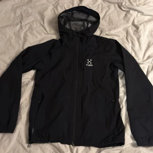 Haglöfs Jacka - Säljer en svart vindjacka från Haglöfs i mycket bra skick. Jackan har en praktisk huva och dragkedja framtill. Perfekt för blåsiga dagar och utomhusaktiviteter. Passar både till vardags och friluftsliv.