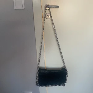 Svart axelväska med kedjedetaljer - Snygg svart axelväska i skinn med kedjedetaljer längs kanterna från Scorett i märket Tiamo. Väskan har en praktisk dragkedja på framsidan och en kedja som axelrem. Fint skick💗