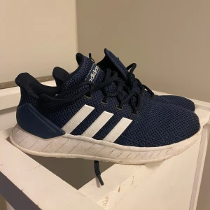 Blå Adidas sneakers - Säljer ett par snygga blå Adidas sneakers i bra skick. De har en stickad ovandel med de klassiska vita ränderna på sidan. Skorna har en bekväm sula och snörning för perfekt passform. Perfekta för både vardag och träning!