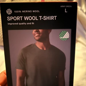 Army Green Sport Wool T-shirt - Säljer en sportig t-shirt i armégrön färg, tillverkad av 100% merinoull. Den har en normal passform och korta ärmar, perfekt för träning eller vardagsbruk. Materialet är mjukt och andas bra, vilket gör den idealisk för både sommar och vår.