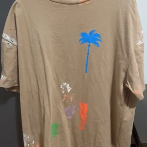 Beige t-shirt från Palm Angels - Säljer en beige t-shirt från Palm Angels med en blå palmtryck på framsidan. T-shirten har korta ärmar och är dekorerad med färgstänk i olika färger. Perfekt för en avslappnad och trendig look. Märkeslogga och copyrighttext på baksidan. Skön och stilren design!