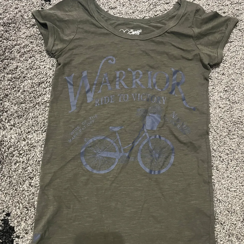 Säljer en olivgrön t-shirt från Sweet Delicatte i storlek S. Den har ett coolt tryck med texten 'Warrior Ride to Victory' och en cykel på framsidan. T-shirten är kortärmad och i bomull, perfekt för en avslappnad stil. Passar bra till jeans eller shorts för en casual look.. T-shirts.