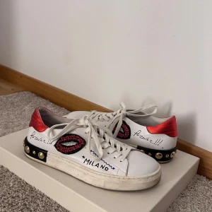 Vita sneakers med röda detaljer/äkta skinn  - Säljer ett par unika vita sneakers med coola röda och svarta detaljer. Skorna har snörning och texten 'Milano' på sidan. De har även dekorativa pärlor och en glittrig läppdesign. Perfekta för att sticka ut i mängden!