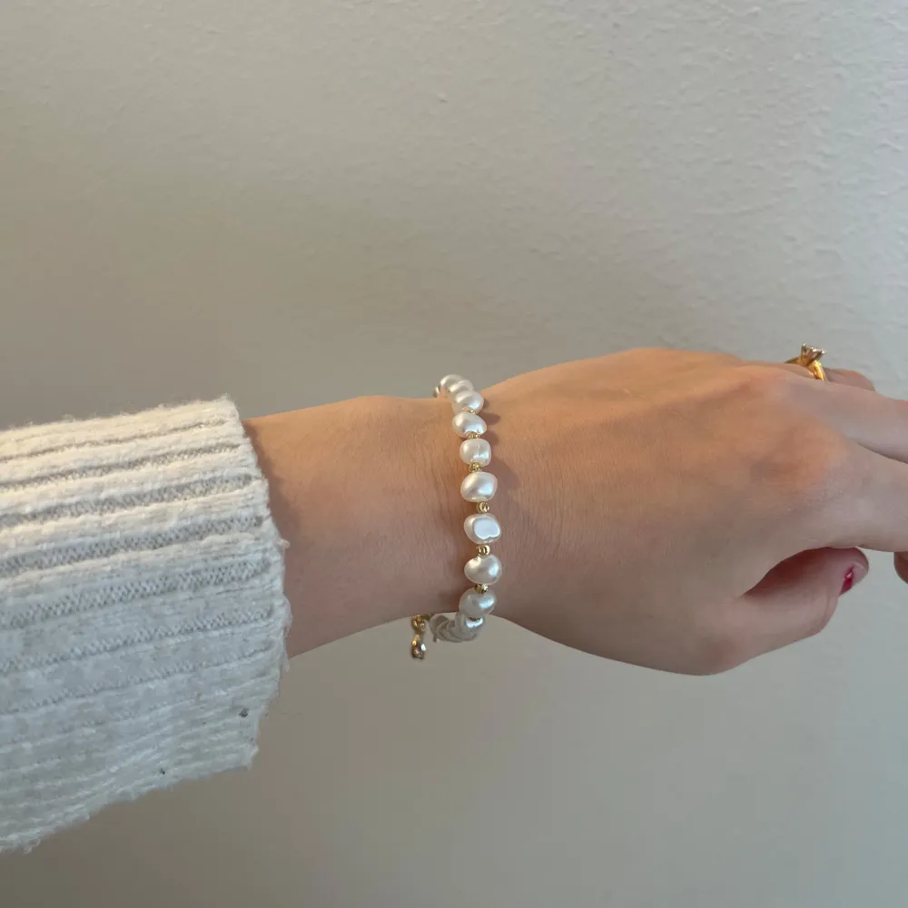 Elegant armband med vita pärlor och guldfärgad låsdetalj. Armbandet har en fin liten hängande dekoration i form av en glittrig insekt. Perfekt för att ge en touch av lyx till din outfit, oavsett om det är för vardag eller fest.. Asusteet.