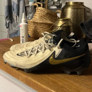 Nike Phantom fotbollsskor - Snygga Nike Phantom fotbollsskor i beige och svart med gulddetaljer. Skorna har snörning och en stilren design som passar perfekt för fotbollsplanen. Nypris 3200 så verkligen värd att köpa till  ett bra pris som man inte får överallt.