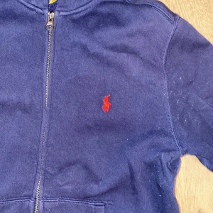 Mörkblå hoodie från Ralph Lauren - Säljer en snygg mörkblå hoodie från Ralph Lauren med dragkedja och röd broderad logga på bröstet. Perfekt för en avslappnad stil skick 9,5-10 inga skador och inga fläckar 