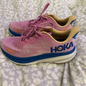 Hoka clifton 9 - Säljer ett par rosa Hoka löparskor (clifton 9) med blå och orange detaljer. Skorna har en bekväm sula och snörning för optimal passform. Perfekta för löpning och träning. Endast använda 1 gång på löpband. Inköpspris 1600kr