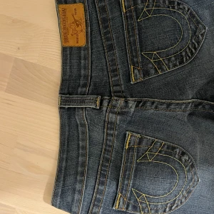 Blå jeans från True Religion - Snygga blå jeans från True Religion med klassisk femficksdesign och kontrastsömmar. De har en lågmidjad passform och är perfekta för en avslappnad stil. Märkets ikoniska logga syns på bakfickorna.