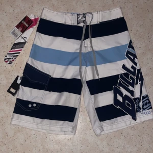 Randiga badshorts från Billabong - Snygga randiga badshorts från Billabong i blått, svart och vitt. De har en cool design med stora bokstäver på sidan och praktisk snörning i midjan. Oanvända med orginalprislappen kvar