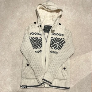 Fodrad Cardigan Jacka - Säljer en beige/vit fodrad cardigan med dragkedja och snyggt mönster i svart och grått på bröstet. Tröjan har en mysig fodrad huva och ribbade muddar. Perfekt för kyliga dagar! Vid minsta fråga eller fundering hör av er🤝